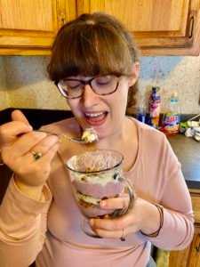 Aimee is holding a No-Bake blackberry cheesecake trifles and holding a spoonful to her mouth.