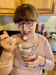 Aimee is holding a No-Bake blackberry cheesecake trifles and holding a spoonful to her mouth.