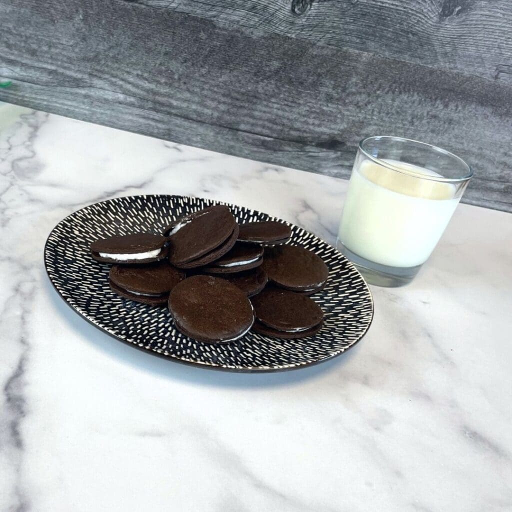 Homemade oreo cookies on a plate with a glass of milk behind the plate on the right side. The plate is dark blue with white lines. The counter is has a white marble pattern and the wall is a dark grey wooden pattern.