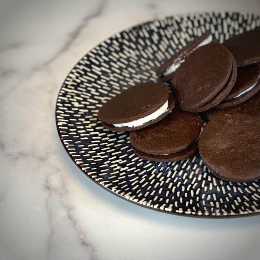 Homemade oreo cookies on a plate with a glass of milk behind the plate on the right side. The plate is dark blue with white lines. The counter is has a white marble pattern and the wall is a dark grey wooden pattern.