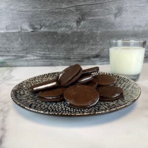 Homemade oreo cookies on a plate with a glass of milk behind the plate on the right side. The plate is dark blue with white lines. The counter is has a white marble pattern and the wall is a dark grey wooden pattern.