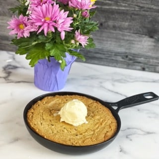  A black cast iron skillet sitting on a white, marble surface and a grey, wooden background.  Behind the skillet is a bouquet of pink flowers.  In the skillet is a large peanut butter cookies with bacon and a scoop of vanilla iced cream on top.